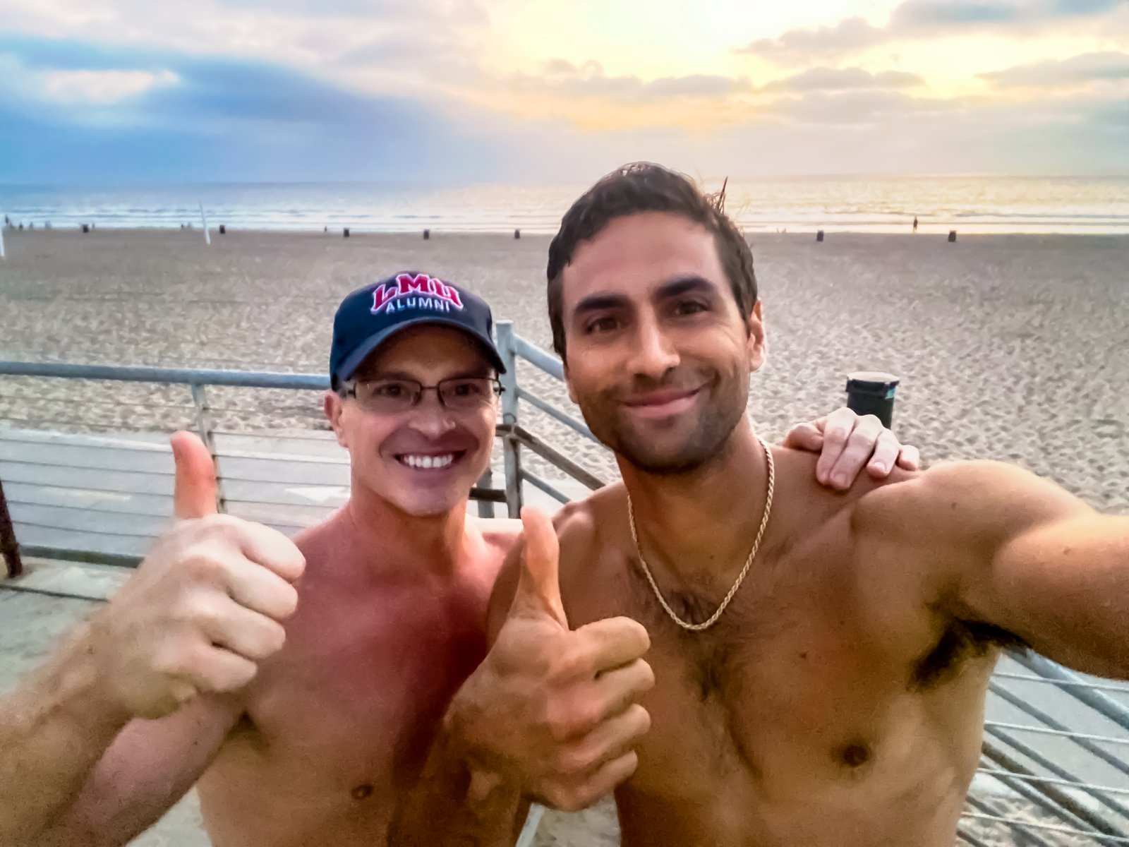 McKay Stewart in an LMU Alumni cap and Ashton Moio, shirtless, giving thumbs up on the boardwalk at El Porto beach at sunset
