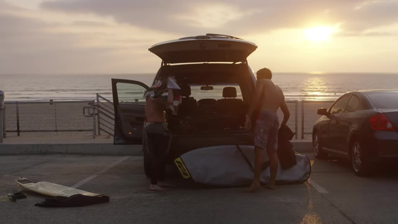 Two men changing out of wetsuits behind an open SUV trunk in the El Porto lot at sunset, surfboard on the ground