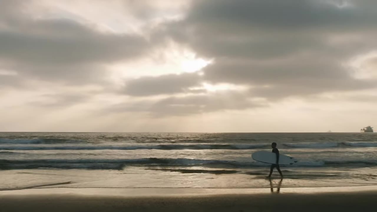 A lone surfer walks along the waterline at El Porto under a cloudy sunset, opening shot of Itchy