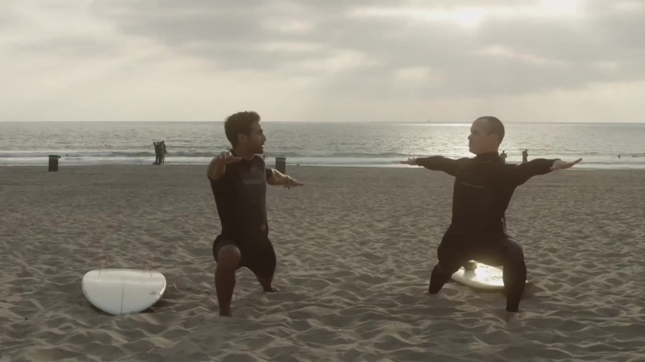 McKay Stewart and Ashton Moio in wetsuits doing warrior-pose stretches on the sand with surfboards beside them
