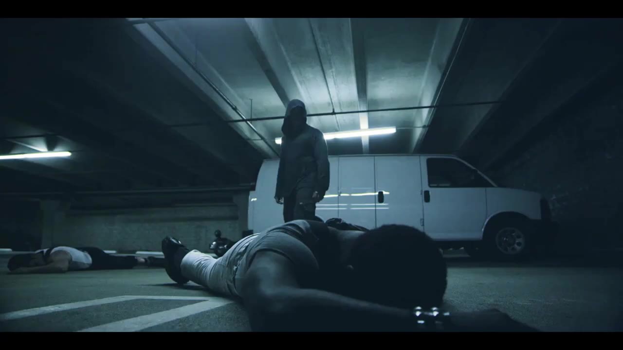 The hooded figure stands over two men lying on the garage floor, a white van behind him